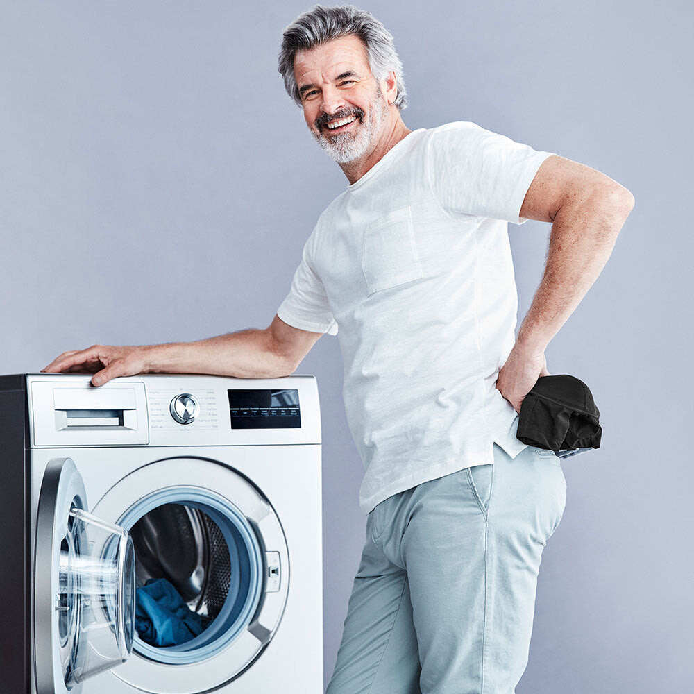 Homme avec une paire de boxers noirs lavables pour incontinence dans sa main, souriant et s'appuyant sur une machine à laver à chargement frontal sur un fond gris.