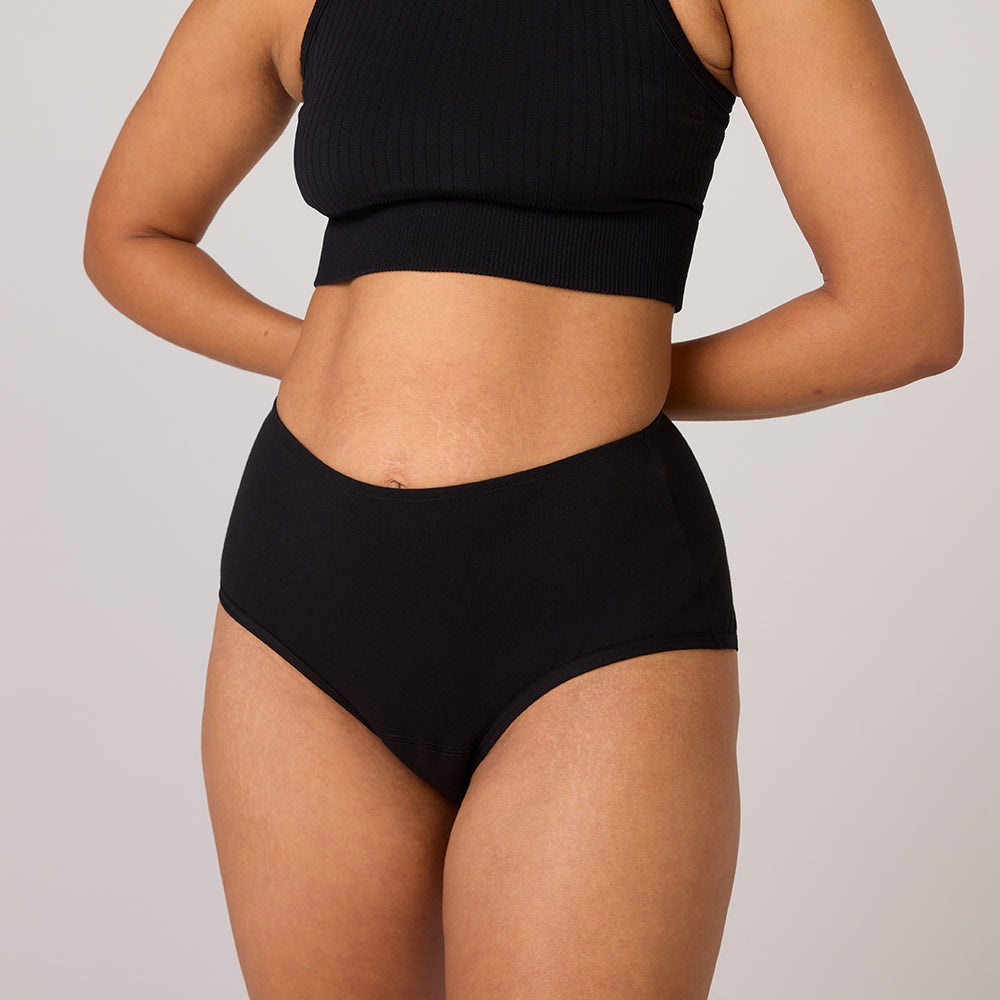 Personne portant un crop top noir côtelé et un sous-vêtement noir taille haute TENA en coton biologique, conçu pour l'incontinence légère, assurant confort et protection.
