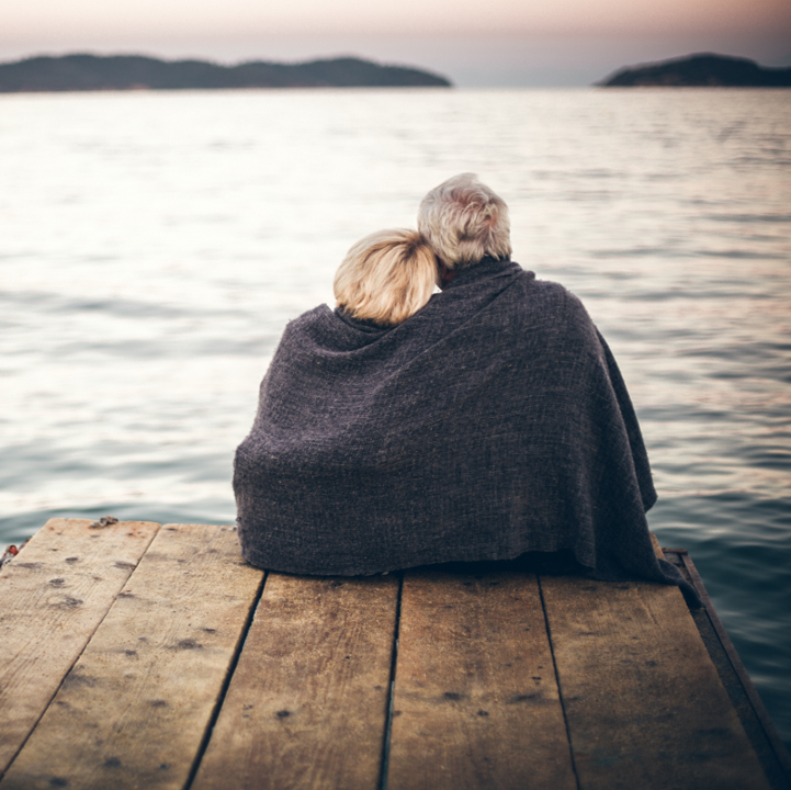 Un homme et une femme sont assis sur une jetée avec une couverture autour d'eux, regardant vers l'océan avec des îles au loin. La femme a sa tête sur l'épaule de l'homme.